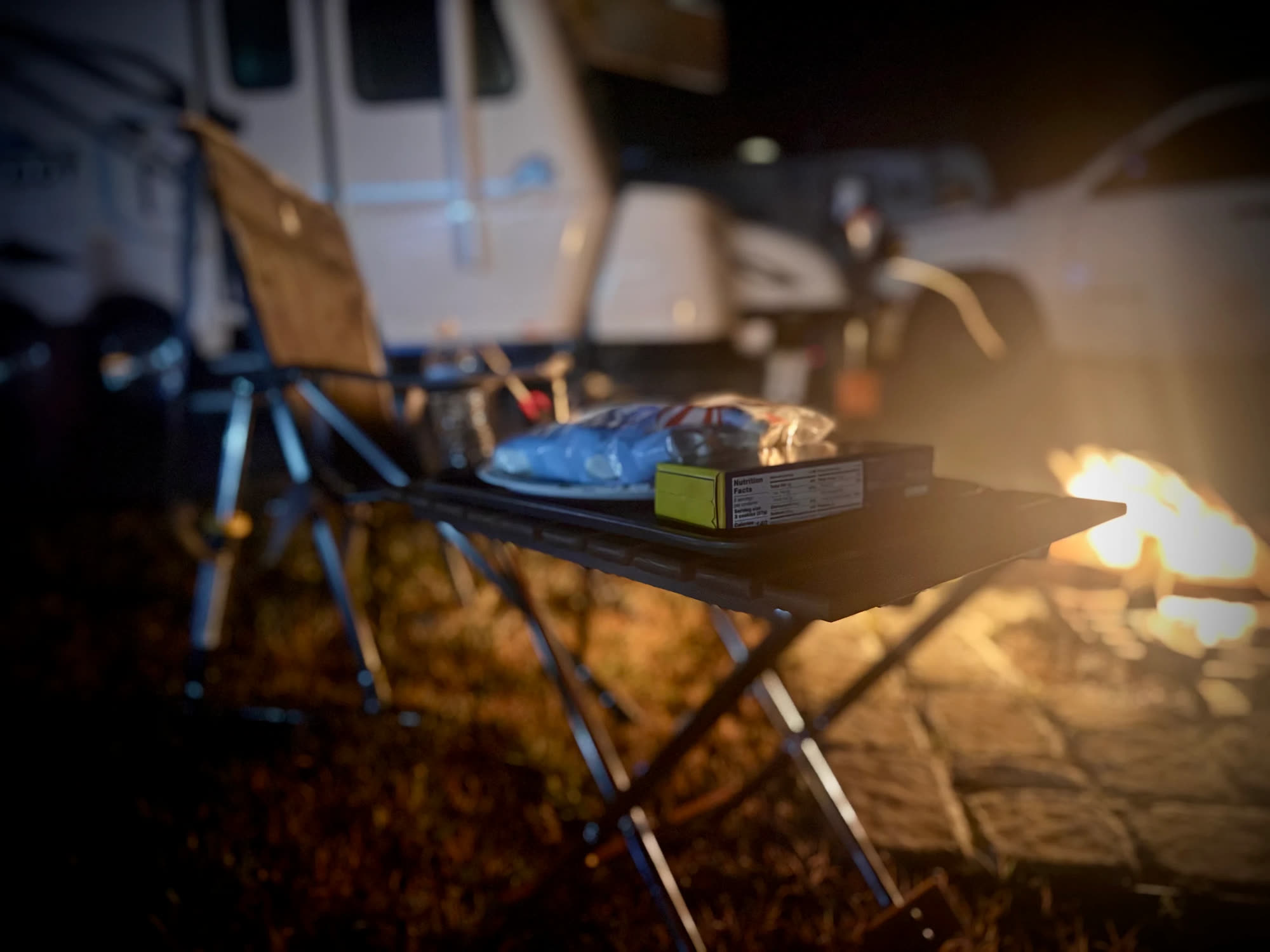 Marshmallows and a camp chair beside a campfire at night, with an RV parked behind the site